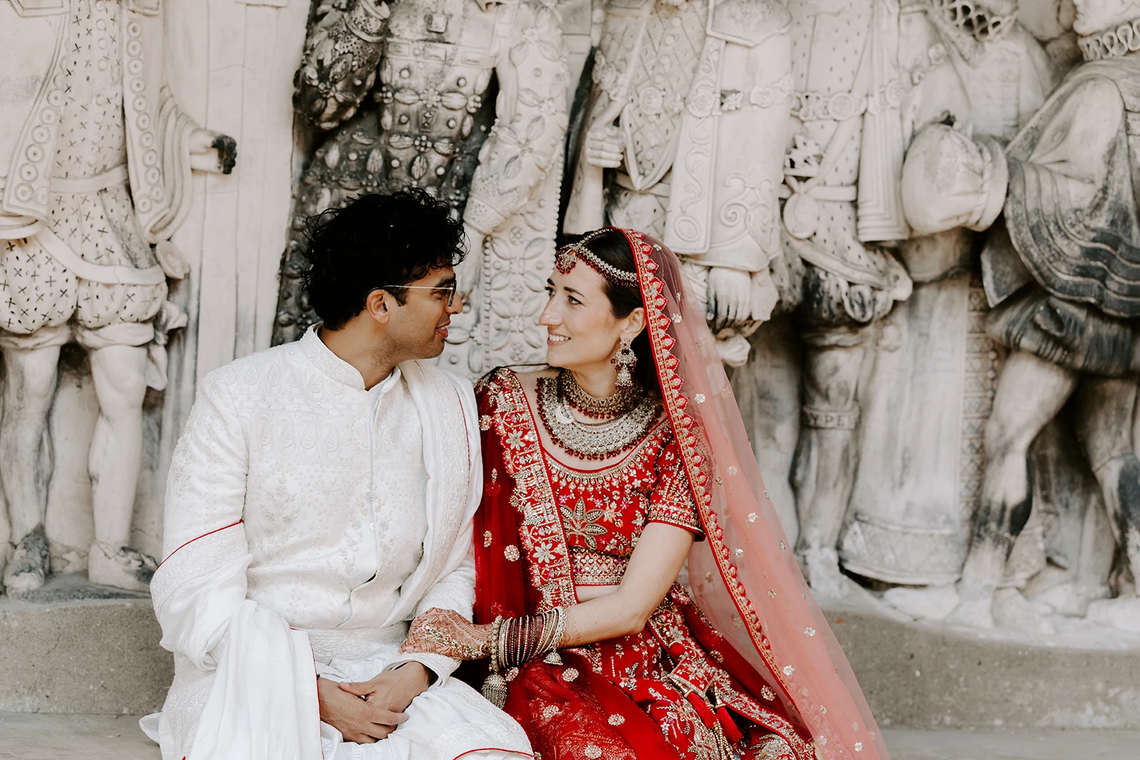 A bride and groom sitting together in front of a stone wall featuring large carved figures of people in historical clothing. A beautiful indian english wedding.