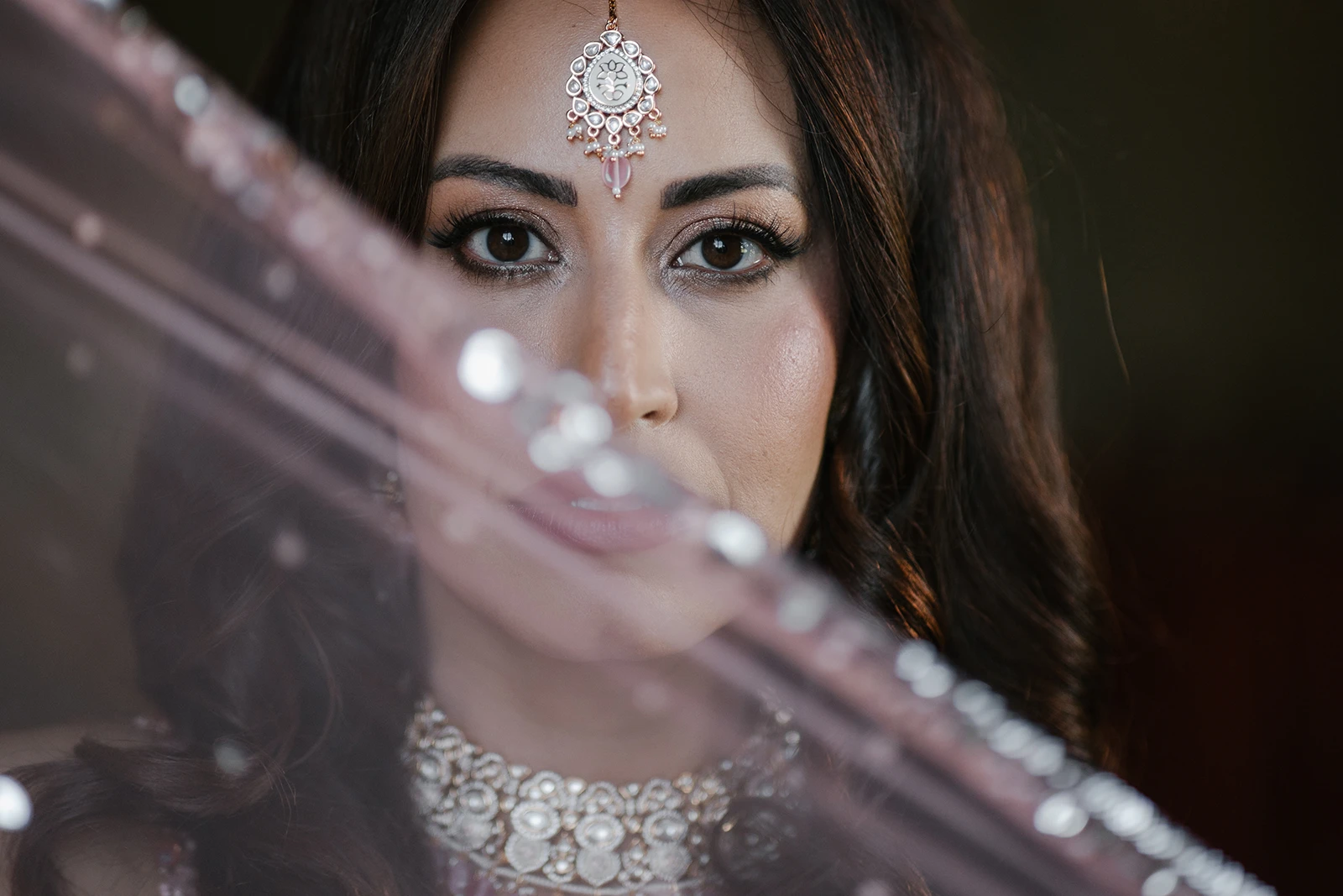 A close-up portrait of a bride wearing a traditional headpiece and necklace looking directly at the camera through a sheer pink veil.