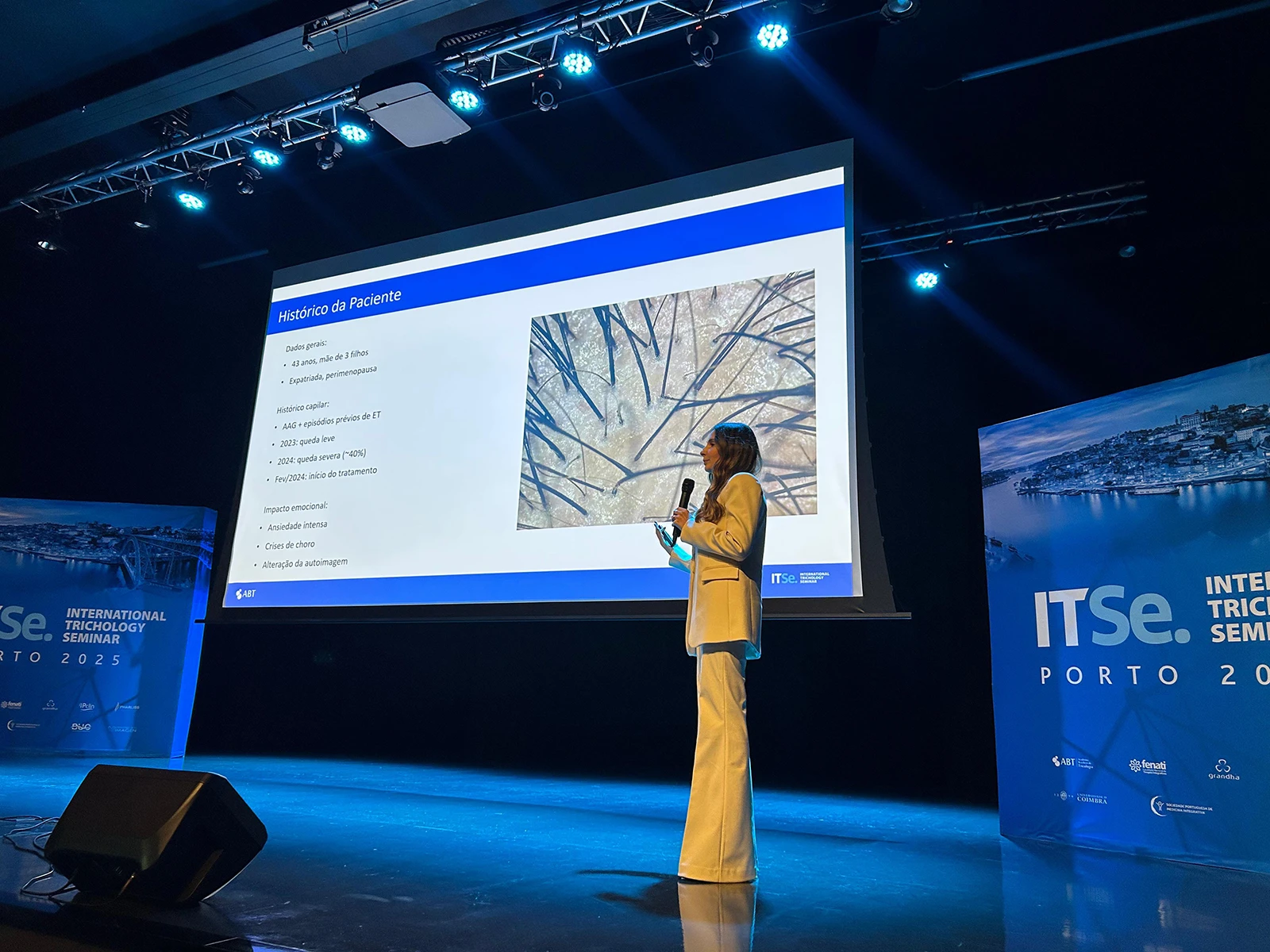 Izabella Bordignon in a white pantsuit presents a case study slide featuring a microscopic hair analysis on a large stage screen at a medical conference.